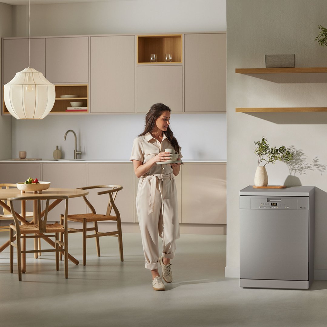 Woman holding dishes, walking past a freestanding dishwasher in a modern kitchen