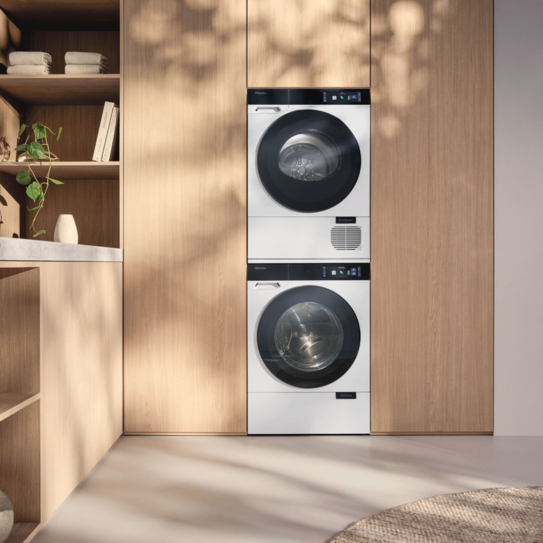 Two Miele dryers stacked in a light wood laundry room with shelves