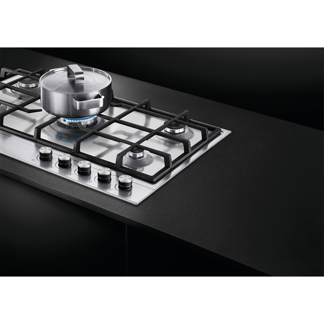 Stainless steel gas cooktop with a pot on a lit burner in a dark kitchen