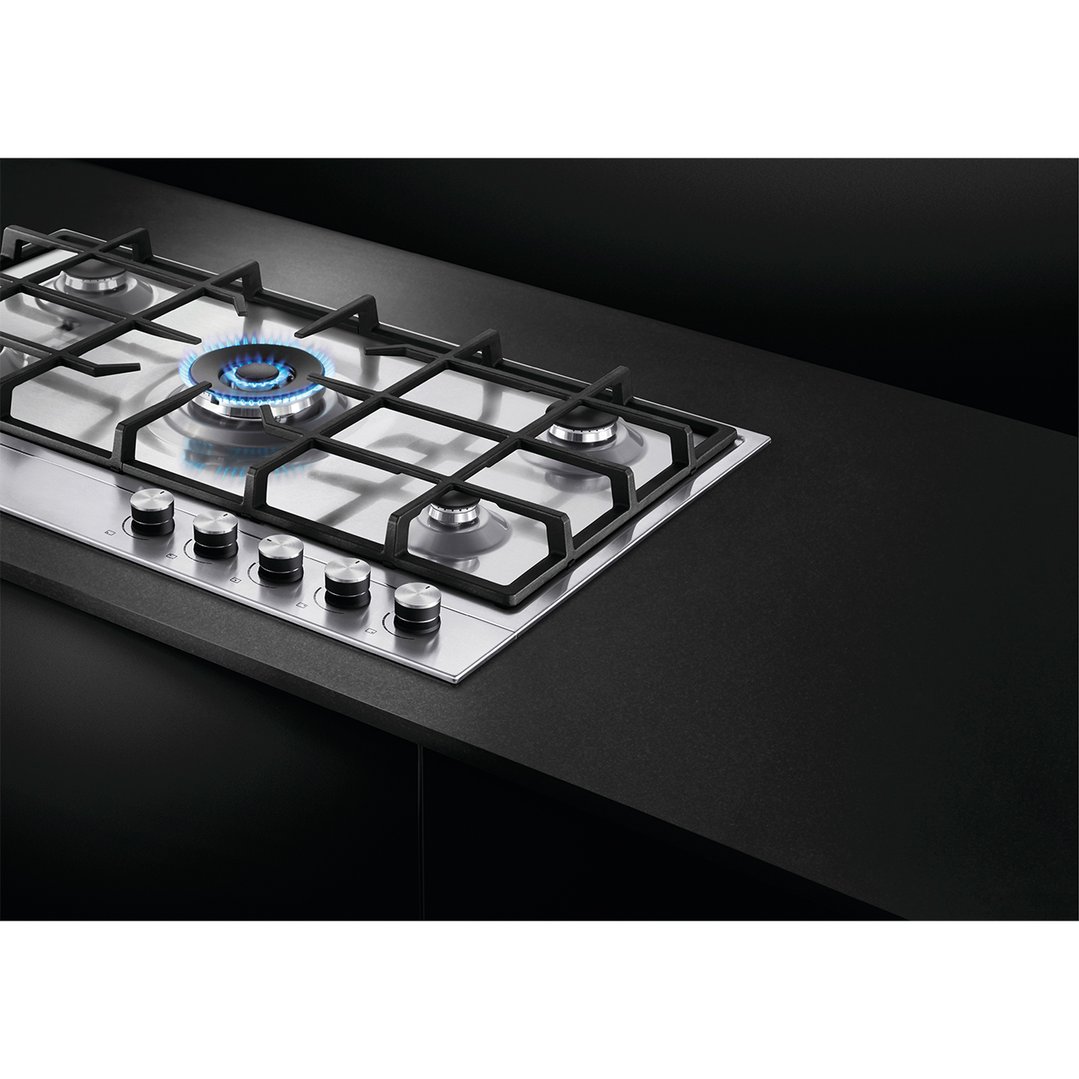 Stainless steel gas cooktop with five burners, one lit, on a black countertop
