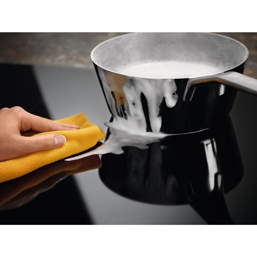 Hand wiping spilled milk from a black induction cooktop with a yellow cloth