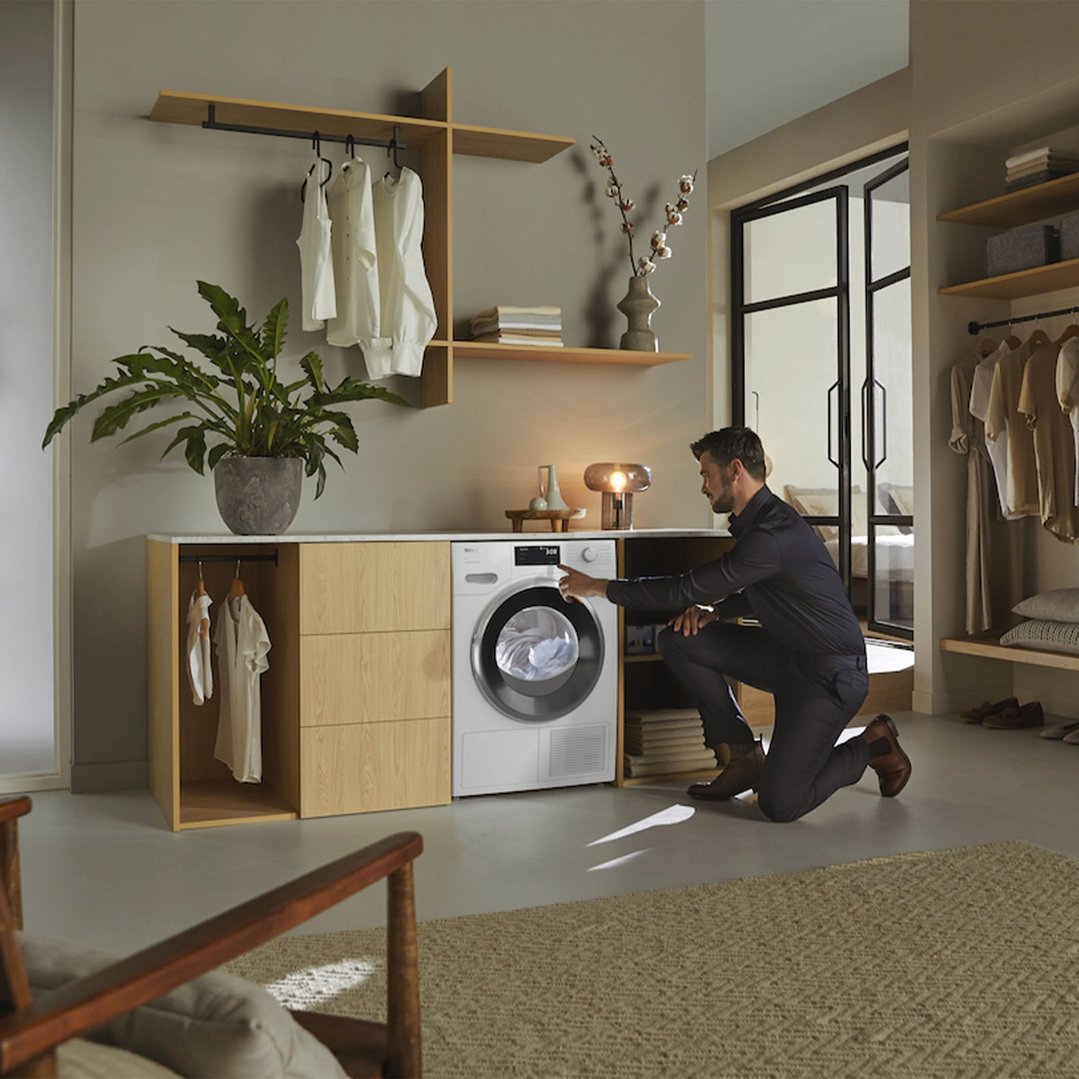 Man kneeling to operate a Miele heat pump dryer in a styled laundry room