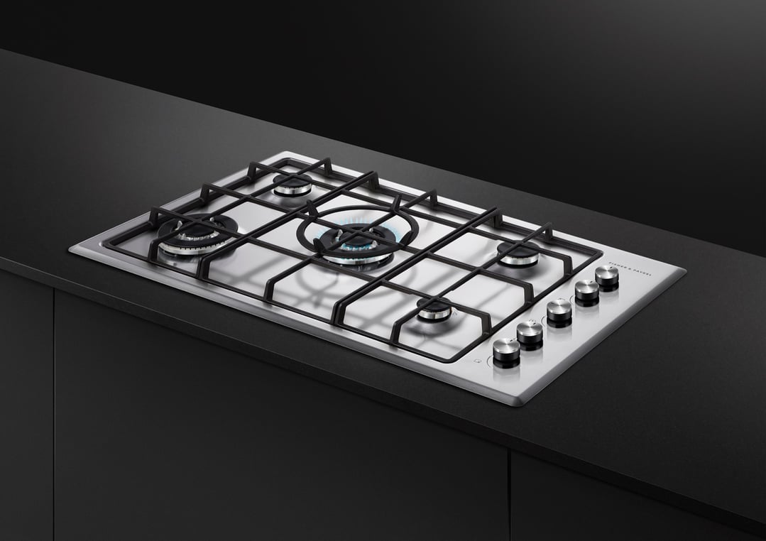 Stainless steel gas cooktop with five burners, one lit, on a dark countertop