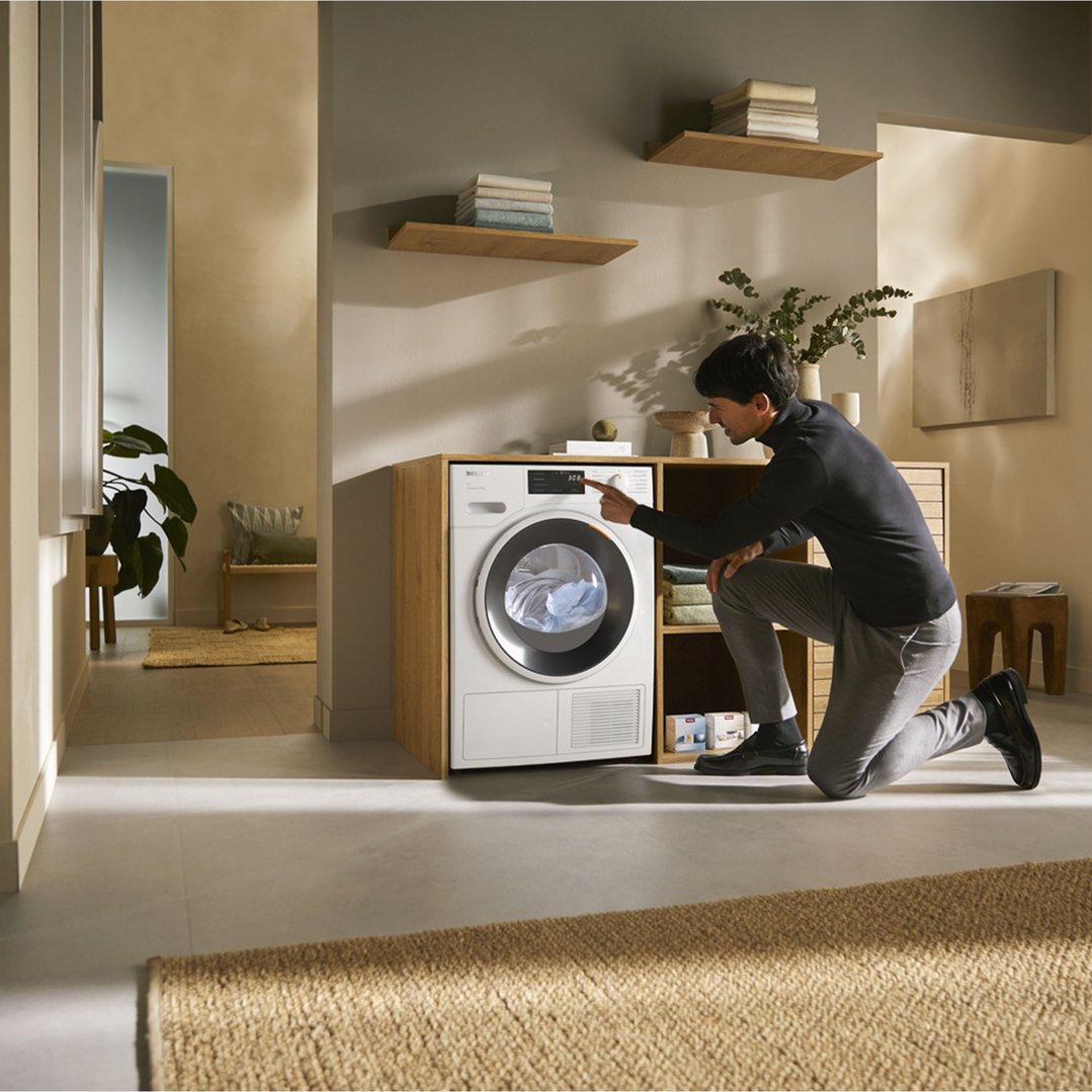 Man kneeling, operating a white heat pump dryer in a modern laundry room