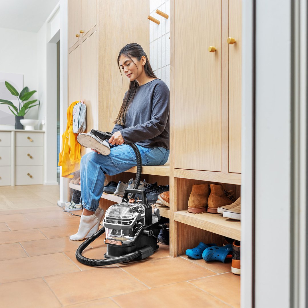 Woman cleaning a shoe with a BISSELL SpotClean HydroSteam vacuum cleaner in a mudroom