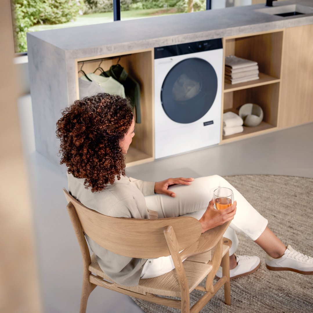 Woman sitting in a chair, looking at a Miele heat pump dryer in a laundry room