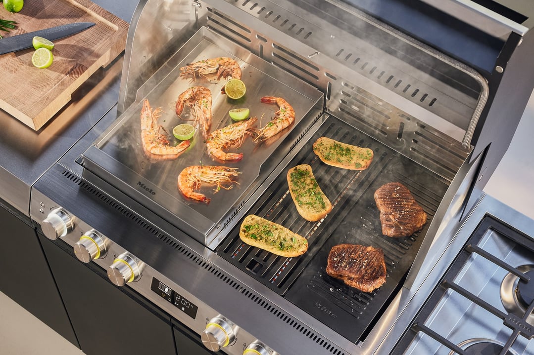 Shrimp, bread, and steaks cooking on a Miele outdoor BBQ griddle plate