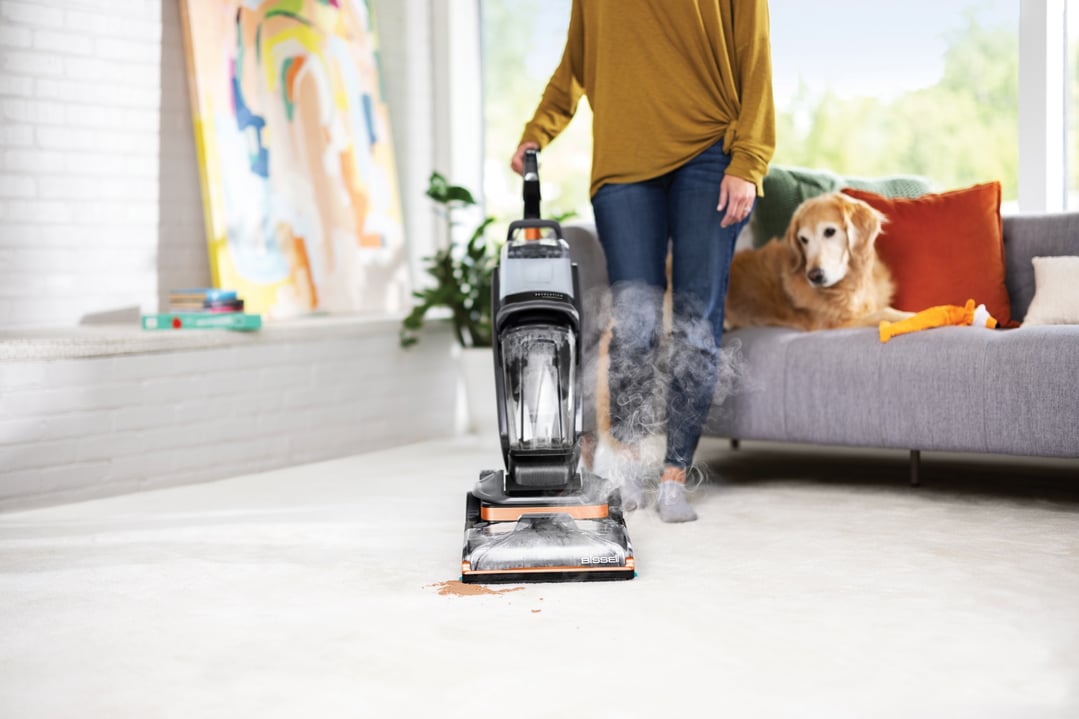Woman using a BISSELL Revolution HydroSteam vacuum cleaner on a carpet with a dog nearby