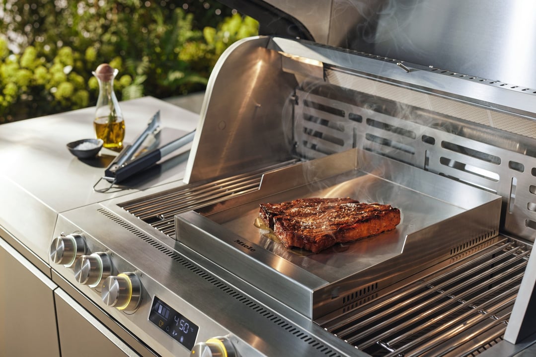 A Miele outdoor BBQ griddle plate with a steak cooking on a grill
